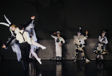 5 dancers are onstage in black and white costumes. In the foreground a dancer with stiff limbs is being carried upstage like a doll by another dancer. The dancers in the background pose stiffly with blank expressions. Photo by Yael Malka.