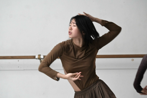 Chi is in motion, dancing with one arm raised and the other in front, her eyes closed as she immerses herself in the movement. In the background is a dance studio. She has shoulder-length black hair and is wearing a brown long-sleeve top and matching brown pants.