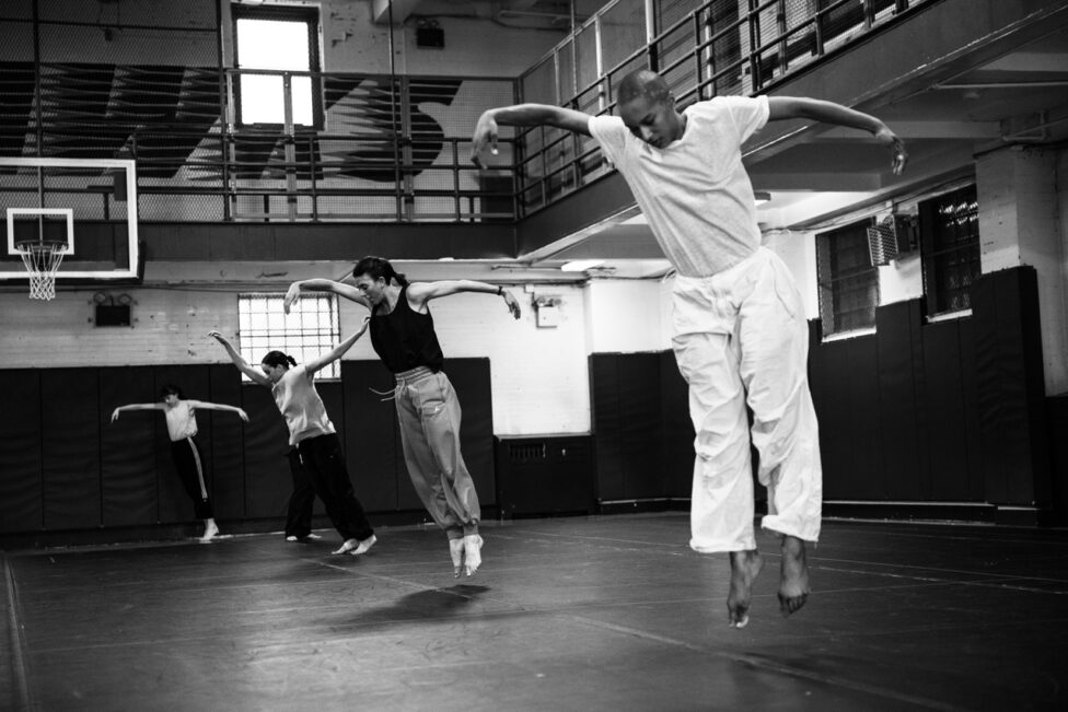 Black and white image of four figures in a mid-air jump, arms arched and faces pointed toward the floor. Photo credit to Maria Baranova.