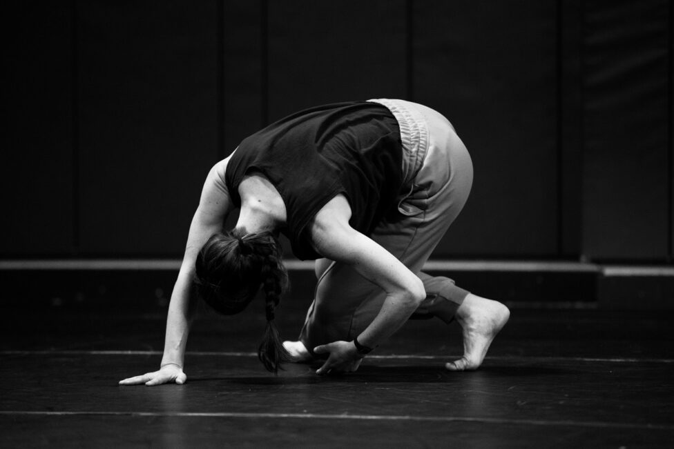 Cecily Campbell presses one hand into the ground and folds inward, the other hand lightly touching the floor. Photo credit to Maria Baranova.