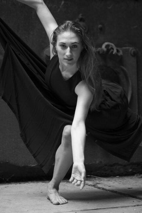 Black and white portrait of dancer in a long black dress balancing on one leg, looking into the camera and extending her left arm down and forward. Photo by Patrick Lanham.