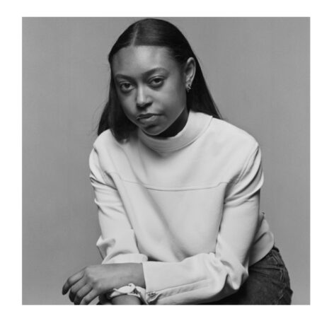 This black and white film photograph shows the upper body of Storm Stokes, a young African American woman with round eyes, dark, straightened hair, wearing an off-white blouse and serious expression.