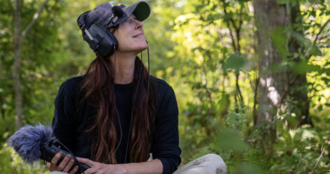 A photo of a person with long dark hair wearing a long sleeved black crew neck sweater and light green pants. This is moira. They are looking up to the left with a big grin on their face while holding an audio recorder in their hands. On moira’s head is a navy baseball cap with a colorful patch on it. A small, furry microphone is clipped to the cap’s brim and large black headphones cover moira’s ears. They are pictured from the waist up, sitting cross legged in a densely green leafed wetland. In the background, bright sunlight dapples across a large tree trunk on the right. The dappled sunlight becomes a glowing trail of bright yellow greens moving deeper into the trees. Photo Credit: Frank DiNardi