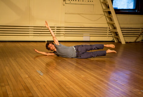 A man in a grey shirt and blue striped pants is laying on the floor on his side with both arms, head and leg reaching off the floor. Photo by Peter Raper.