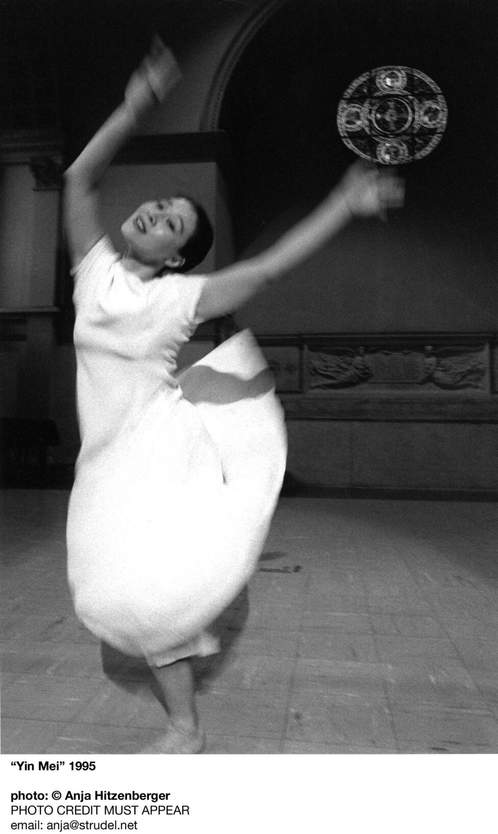 A photo of Yin Mei performing at the Judson Church. Yin balances on one leg while her arms wave above her. Her dress billows behind her in motion. Photo by Anja Hitzenberger.