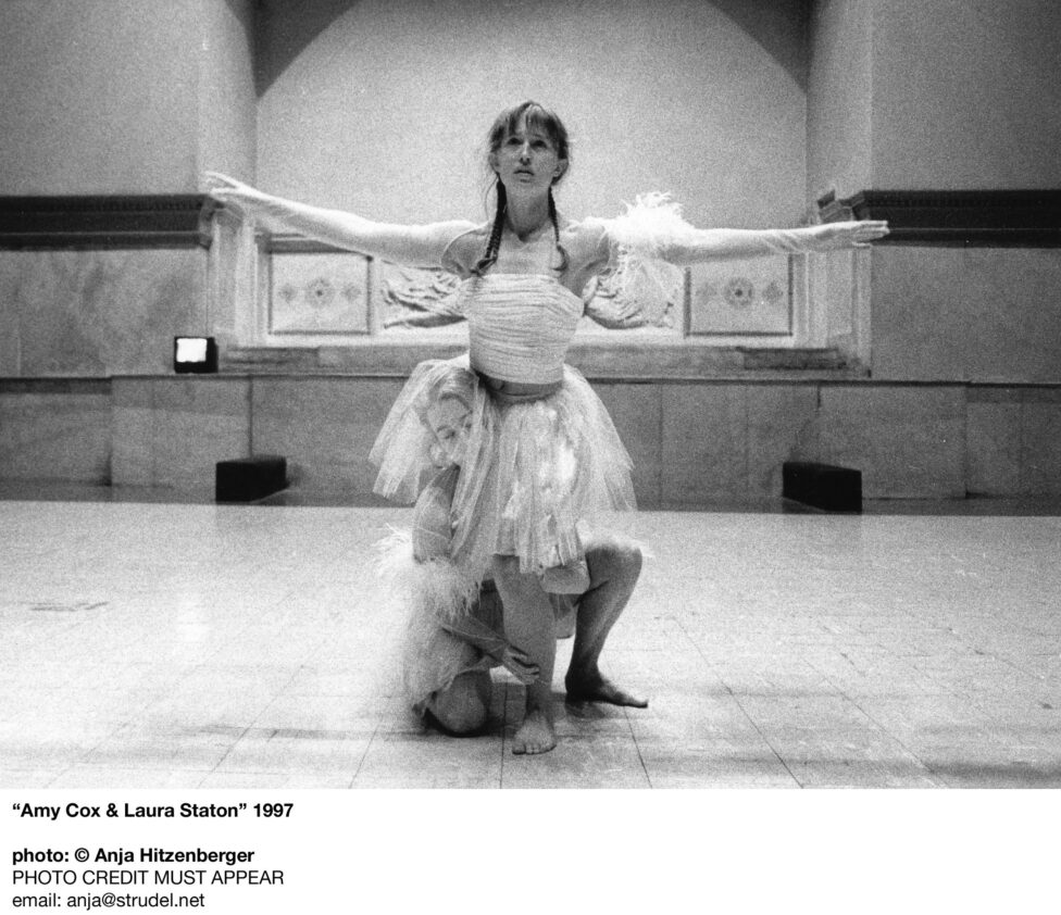 Photo of Amy Cox and Laura Staton performing at the Judson Church. One dancer crouches on the floor supporting the other who straddles their shoulder, looks straight ahead and holds their arms to the side in a T formation. The dancers wear gauzy skirts. Photo by Anja Hitzenberger.