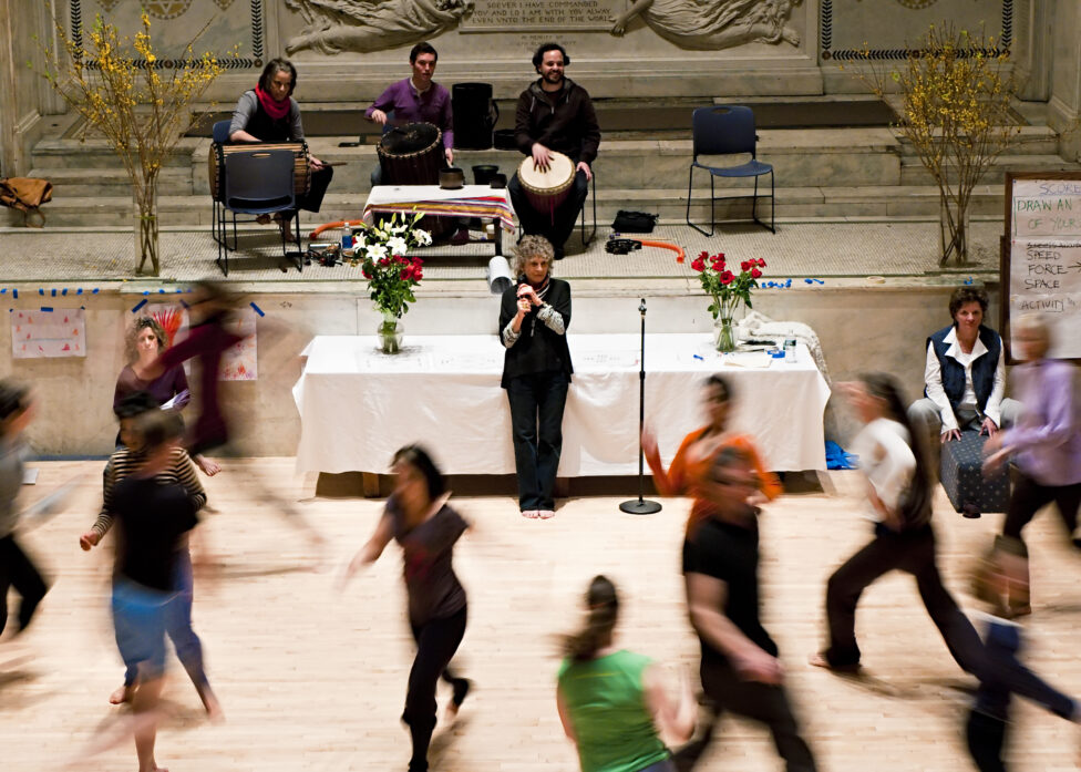 Anna Halprin stands center in front of a table with a cloth and vases of flowers. Blurry dancers move in front of her.