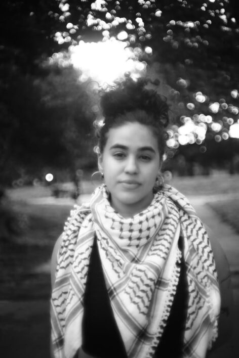 Anaís is standing with a keffiyeh on at the park in a black and white image in their hoop earrings