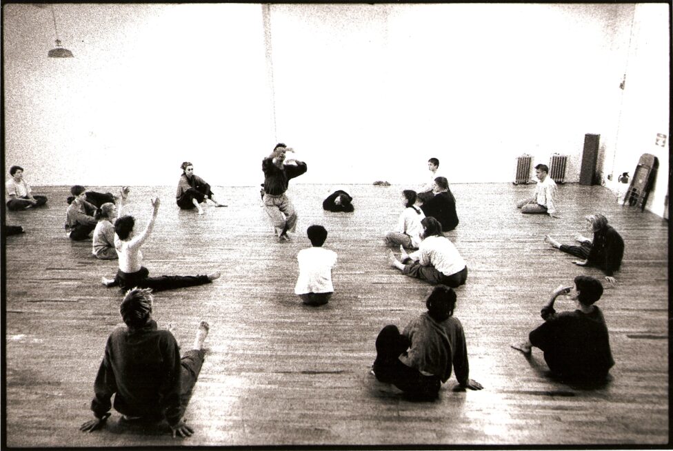 Sepia toned image of dancers sitting and observing another dancer in the center of a studio with hardwood floor. From the MR Archives. Workshop 1993 with David Zambrano photo by Anja Hitzenberger.