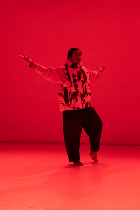 A brown femme with short curly black hair is in a blue and white puffer and sparkly baggy jeans with her arms opened to the side with one foot propped in a demi releve and the other foot firmly planted on the floor. She is saturated in a deep red light that envelops the whole space.Photo by Elyse Mertz courtesy of Center for Performance Research.