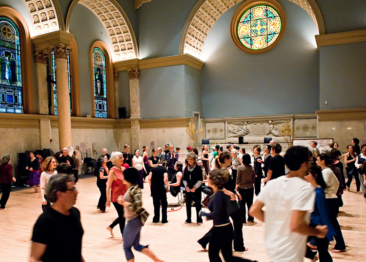 From the MR Archives, 2010. Photo by Ian Douglas. People gather and move about in spance, Anna Halprin stands in the midst of it all inside an open indoor church.