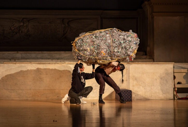 A photo of Earthworks Performing for Movement Research at the Judson Church. Two dancers carry and support a large bundle of plastic bottles. Photo by Rachel Keane.