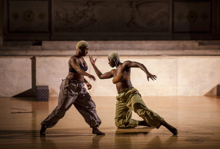 A photo from Stephen Shyness' work for Movement Research at the Judson Church, AoCC curation. Two dancers in motion. One dancer steps forward gesturing with their arm. Another supports their body kneeling on their right side with their left leg extended out. Their arms reach in opposite direction. They both have bleached blonde short cropped hair. Photo by Rachel Keane.