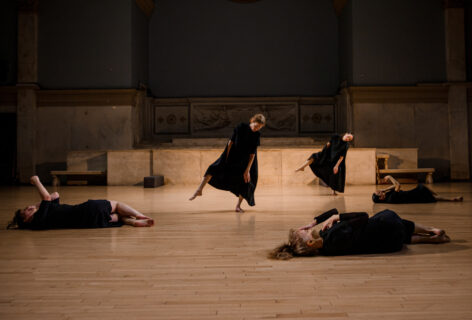 John Jaspers work featuring five dancers dressed in black dresses. Three dancers lay on the floor knees to one side and back flat to the ground while they gesture and touch their hands to their faces. Two dancers in the center balance on one leg with their torsos leaning diagonally off their center. Photo by Rachel Keane.