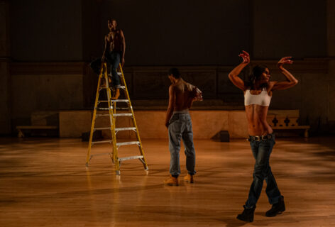 A photo of ms. z tye and two dancers performing. ms. z tye is in motion stepping forwards with both arms raised above their head elbows bent and wrists bent with soft fingers. One dancer faces away from her looking towards a ladder where another dancers sits straddled at the top. Photo by Rachel Keane.