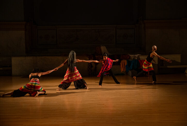 A photo of Ani - Malaya Works piece for Movement Research at the Judson Church. Three dancers in a diagonal line holding on to eachother's hands. They are spread out and reaching. One dancer at the end of the line is on the floor. A fourth dancer moves away from the group. Reaching their arm as they move forwards. All dancers wear multi colored fabric with red yellow and green panels. Photo by Rachel Keane.