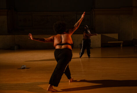 angel shanel edward's work for Movement Research at the Judson Church. Two dancers moving and placed diagonally from each other. In the foreground one dancer lunges away from the camera with arms extended upwards and sideways. Upstage the second dancers stands with one arm swinging across their torso. Photo by Rachel Keane.