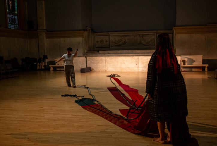 Two dancers stand diagonally facing each other. The dancer in the foreground holds red fabric panels that stretch out across the floor towards the other performer. Photo by Rachel Keane.