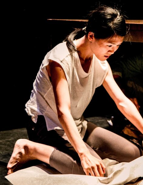 A woman with short dark brown hair pulled into a ponytail wears a baggy sleeveless white top, brown shin-length pants, and bare feet. She is kneeling as she looks down at a diagonal direction in front of her, extending both arms in that direction. Photo by Taso Papadakis