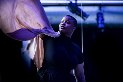 Enya-Kalia Jordan, a Black woman donning a shaved head, looks upward as she gently floats a light peach-colored scarf above her shoulder. She is framed by the background of a dance studio mirror and a gentle light caressing her face. Photo by Ryan S. Brandenberg