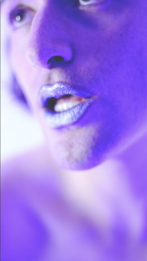 A portion of a face seen close up, with dramatic blue lighting highlighting the individuals pale features, sparkling silver lips, and strong nose. Photo courtesy of the artist.