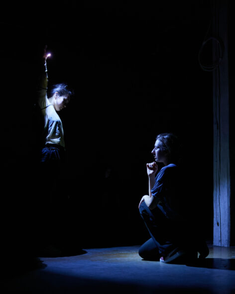 Giordana stands in darkness and stretches an arm high above her head, shining a pool of light onto a cement floor with a flashlight. Agata sits in this circle with eyes closed and her chin resting on her hand. Photo by Allina Yang.