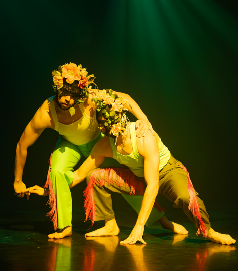 A dancer is crouched on all fours peering into the distance while another dancer stands eract on his lower back. The standing dancer reaches for a rainfall of orange petals falling from the ceiling. Photo by John Landry