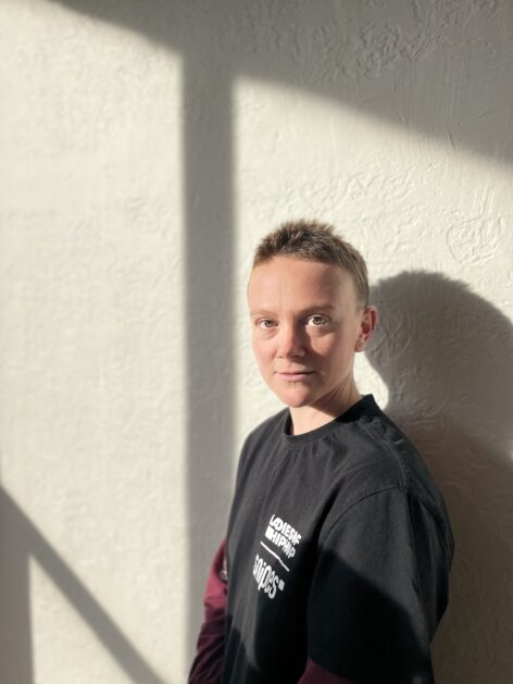 Paris stands facing the camera, visible from the chest up against a white wall with shadows cast. They're wearing a black t-shirt with white lettering, have light brown eyes, and short brown hair. Photo courtesy of the artist.