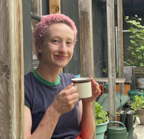 Sam Kann, a white queer person with short pink hair, smiles at the camera holding a tea cup.