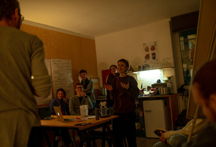 The photo shows a playful performative situation in the kitchen of the Dance Residency Centre in Bratislava. It shows Sonia laughing at her co-performer Daniel. In the background there are spectators and our friends who are observing us with a smile. Photo by Oleksandra Sherhina.