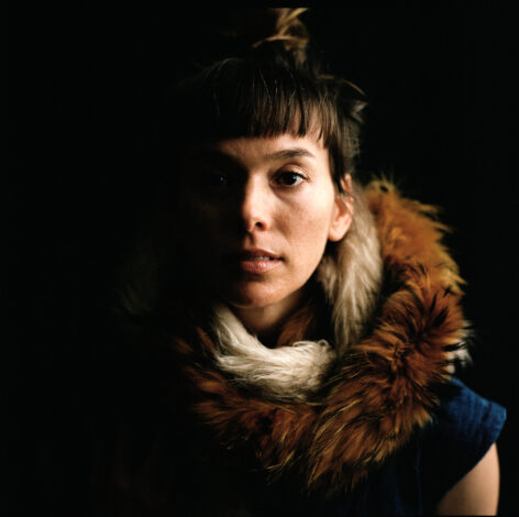 marion looks straight at the camera. One side of her face is lit, the other obscured by shadow. Her brown hair is pulled up on top of her head. She is wearing a hand-me-down denim jumpsuit from Michelle Boulé and a brown fur scarf around her neck. Photo by Maria Baranova