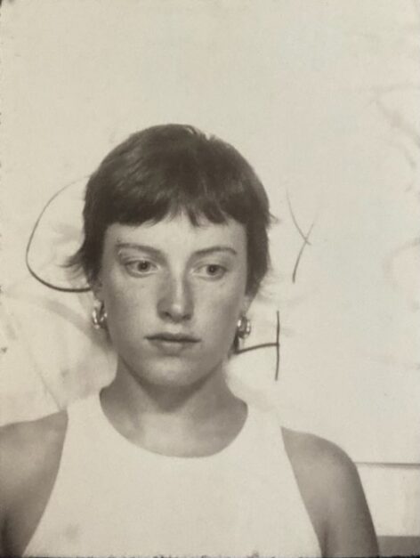 Melle is pictured in a black and white photo booth wearing a white tank top and looking down. She has short brown hair and hoop earrings on.