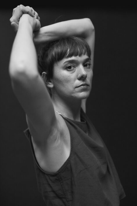 Marion stands in profile view looking at the camera, her hands embraced above her head. Photo by Anna Maynard