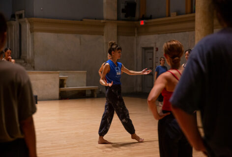 From the MR Archives, 2023 Summer MELT. Joanna Kotze teaches at the Judson Church. She wears a blue top and patterned bottoms. Her arms out and one foot stepping forward. Photo by Rachel Keane.