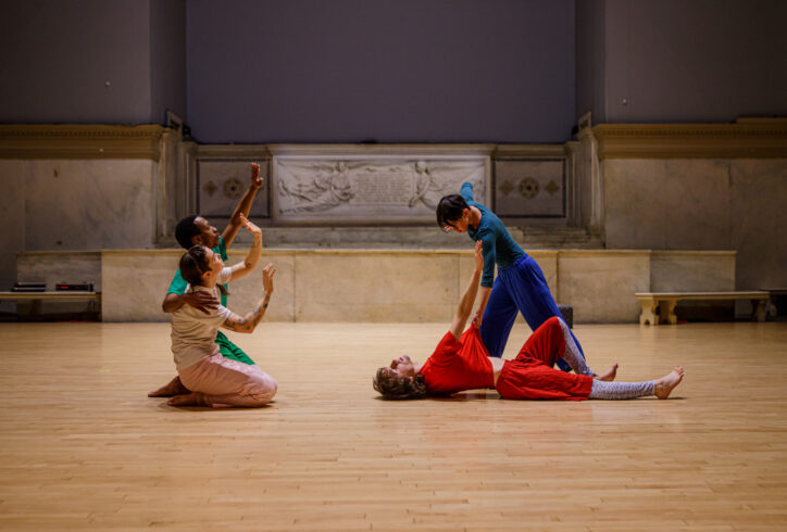 A photo from Jade Manns performance for Movement Research at the Judson Church. Two dancers kneel and gesture with their arms reaching upward and their palms facing out. Two other dancers are engaged in a movement. One dancer wearing red lies on the floor and reaches towards the second dancer wearing blue. The second dancer leans over pulling the first dancers shirt. Photo by Rachel Keane