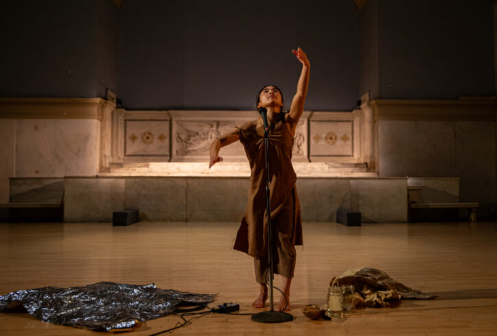 A photo of Marie Lloyd Paspe's performance for Movement Research at the Judson Church. Marie reaches upwards with one arm, their face follows their gaze. They wear a brown dress and stand in front of a microphone. At their feet there is a sheet of metallic material, glass jars with water and crumpled brown paper. Photo by Rachel Keane