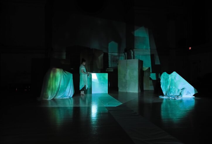 A photo from alys longley with Proyecto Undular-Undular Projects' performance for Movement Research at the Judson Church. A dancer stands amongst many boxes and draped fabric in a dark environment. Blue green light washes over the scene. Photo by...