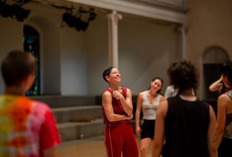 Jennifer a white woman with a black bandana wearing red pants and a red tank top is smiling large / laughing and gathered with a group of MELT participants at St. Marks Church in the Bowery. Photo by Rachel Keane.