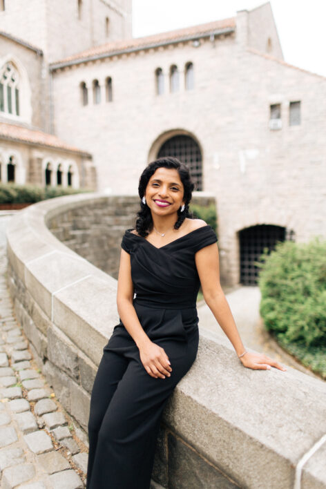 Aparna Shankar vertical headshot: Aparna is wearing a black jumpsuit and bright purple lipstick sitting on a stone ledge. Photo by Blue Spark Photography.