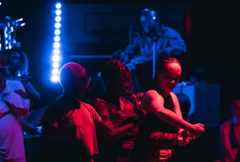 Three Black women exchanging gestures in movement side by side with various colors. A Black man in the background offering percussion as music accompaniment immersed in blue light. Photo by Henry Creel.