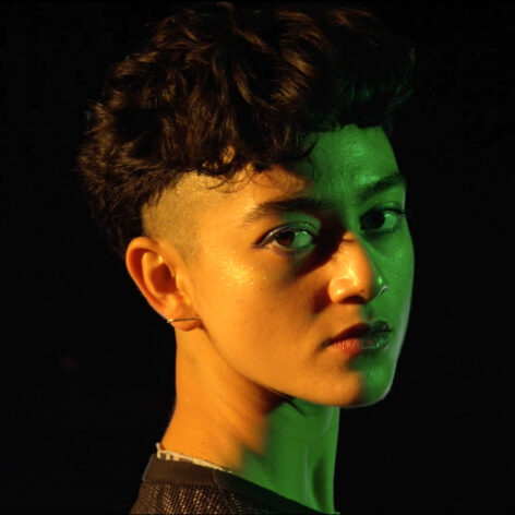 Close up of Luna Beller-Tadiar's face, half lit in orange and half in green. She has short curly black hair with shaved sides and light brown skin that looks orange in the light. They look at the camera. Photo courtesy of the artist.