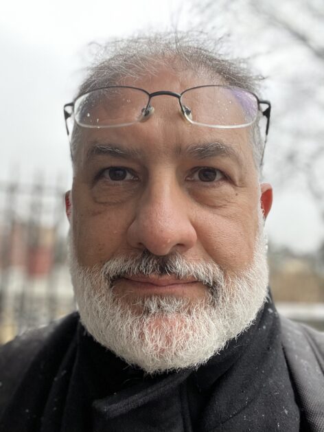 A man with a white beard and reading glasses on his forehead. He looks at the camera. Photo by Hossein Aleyasin (Selfie)