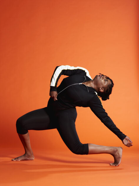 A Black Woman dressed in black and white, with dark hair wrapped in a bun, displaying profiled of full bodied motion very low to ground in a back-bend position. Photo by Canaan Mattson.