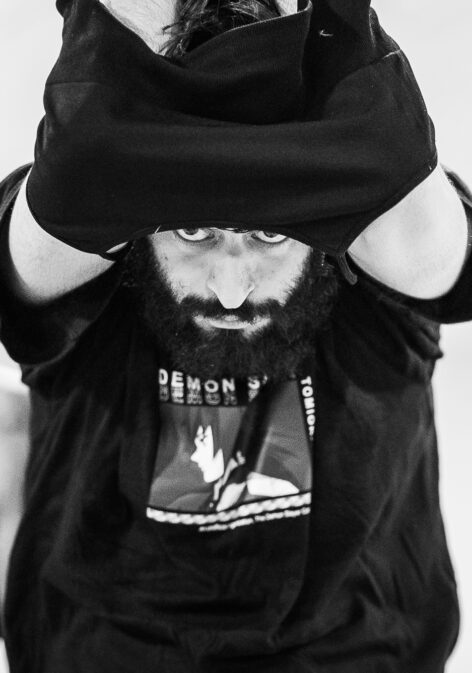 A photo of Mohammadreza Akrami. He peaks put from under a a shirt he is caught in. His arms extend over his head and his face looks directly into the camera. They have a beard and wear a top with an anime character. Photo by Arnaud Beelen