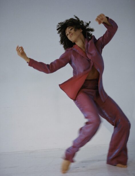 A light skin black woman with short curly hair wears an iridescent pink outfit. She lifts her hands towards the sky and gazes to the right. Photo by Nigel Hosang.
