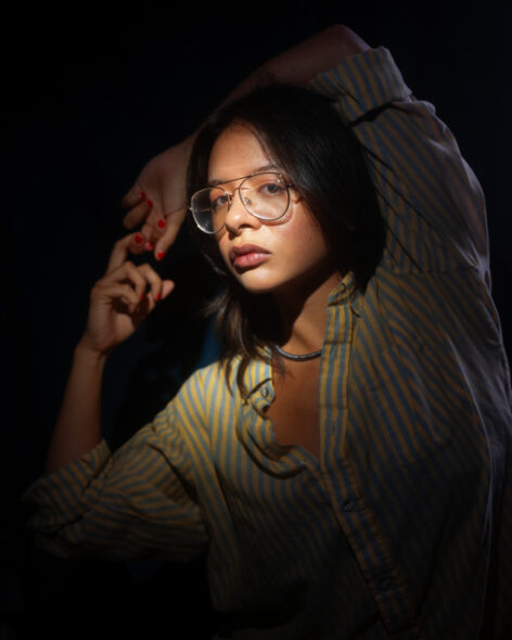 A woman with brown hair, brown skin, and glasses poses in a dramatic light. She is wearing a yellow and grey striped shirt with her arms outstretched to her right. Photo by James Gentile