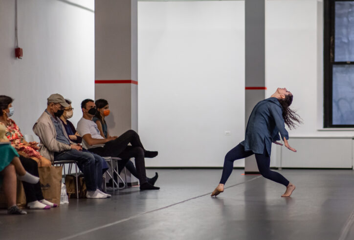 From the MR Archives, 2022. June, 7. Open Performance Photo by Rachel Keane. Audience masked sit in an open studio, once dancer arches back with arms down slightly lunging forward on a forced arch. Their focus is towards the ceiling.