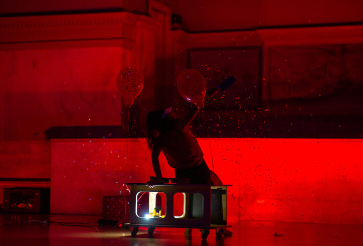 From the MR Archives. Martita Abril performs at Judson Church in a red light. They sit on a bench with wheels that have 2 balloons tied to it and a work light tucked in. She looks off the right with a butcher knife in her hand and confetti falls around her. Photo by Rachel Keane.