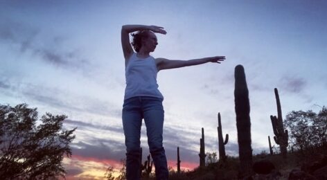 A photo of Nicole Touzien standing in front of a sunset. There are cacti around her and she reaches her arms over her head to one side. Photo courtesy of the artist