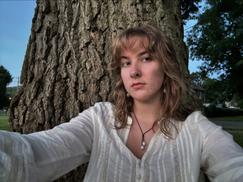 A woman with wavy blonde hair, blue eyes, and a silver nose ring sits against the trunk of an oak tree. They wear a white blouse with a pearl necklace and pearl earrings. She bears a neutral expression.